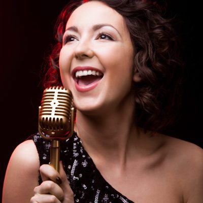 Beautiful young female singer in sparkling black evening dress singing holding golden vintage microphone, during live musical performance